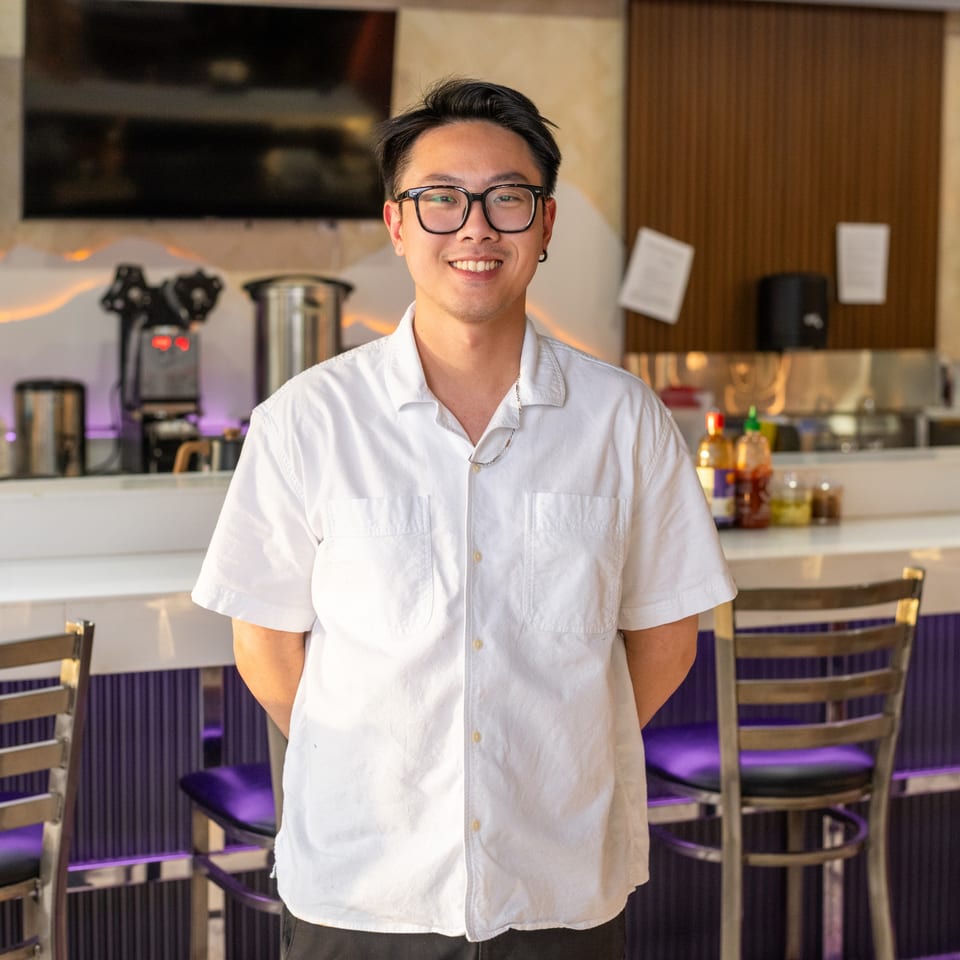 Smiling Asian man in glasses and white shirt standing in front of a restaurant bar.