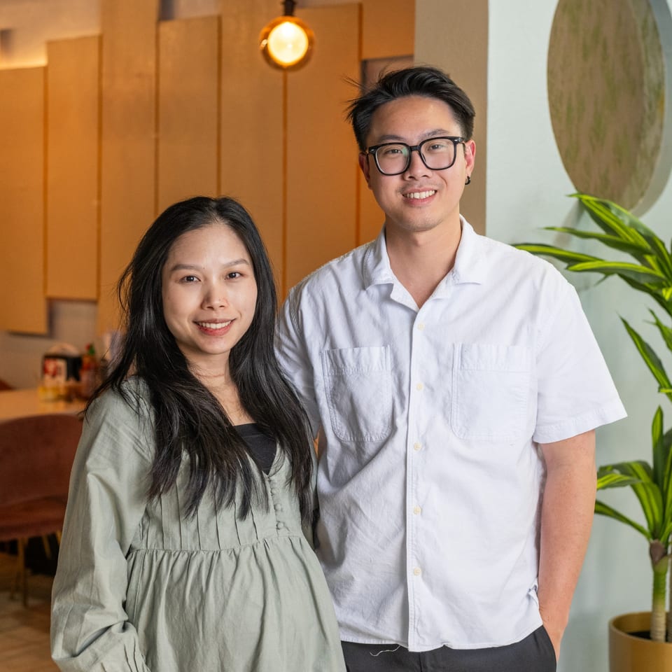 Two smiling people, a woman and a man, stand together in a modern, warmly lit interior.
