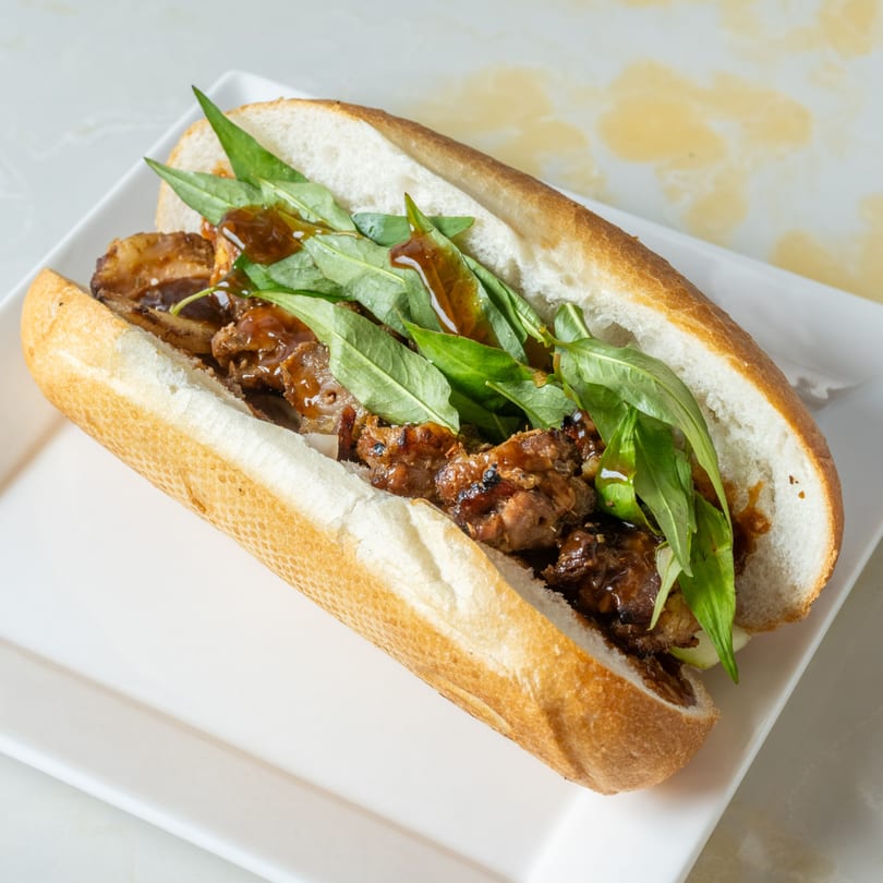 A Vietnamese Banh Mi sandwich with savory meat, fresh greens, and a rich sauce on a white plate.